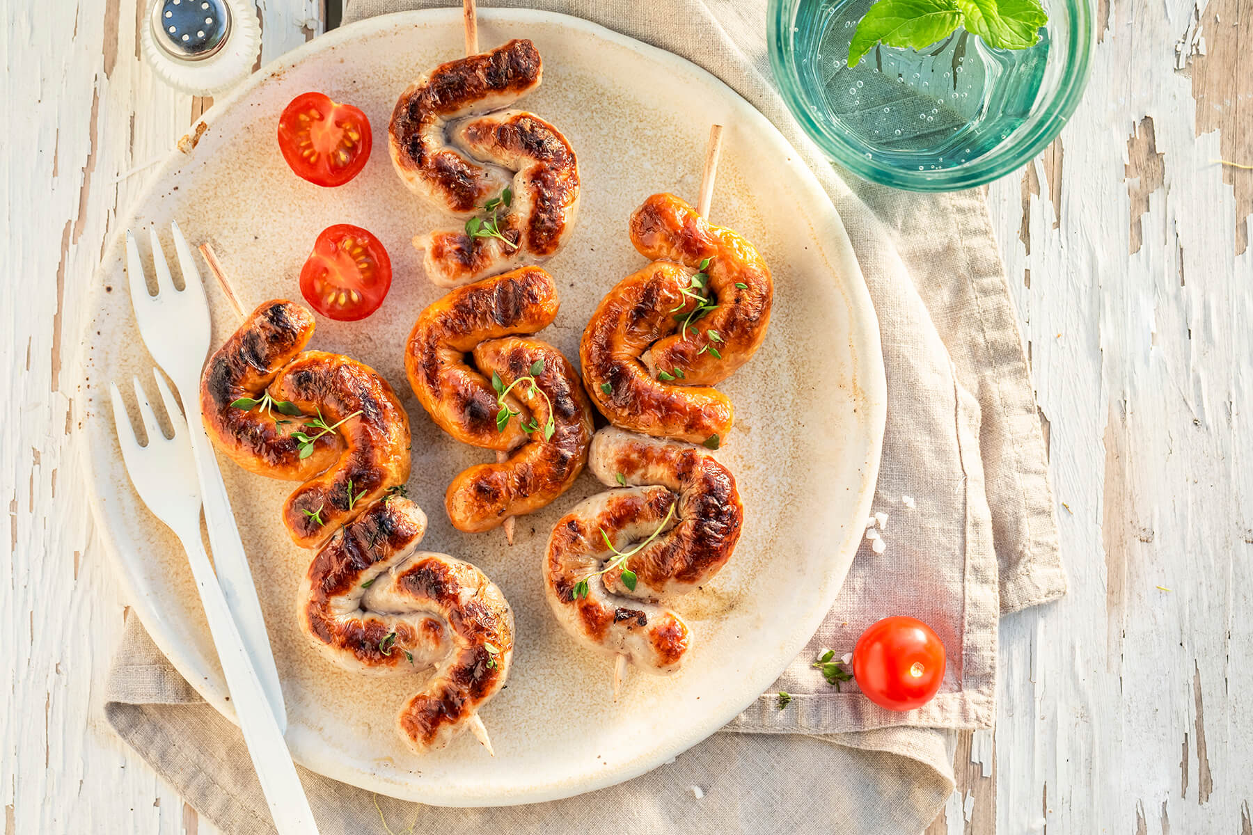 Gefaltete Bratwurst am Holzspieß auf einem Teller mit halbierten Kirschtomaten, Thymian und einem Glas Wasser