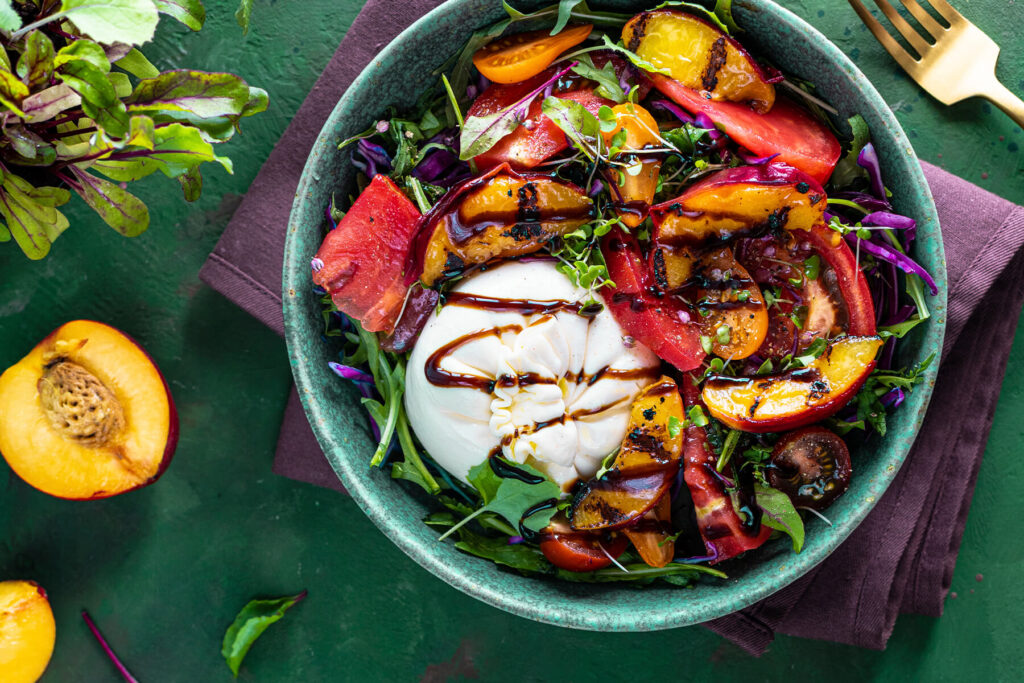 Sommersalat mit Burrata und gegrillten Nektarinen von oben in einer türkisfarbenen Keramikschale: eine ganze weiße Burrata mit dunklen Balsamico-Bändern, darum Nektarinenspalten mit Röststreifen, rote Tomatenspalten, orange und dunkle Kirschtomaten, fein gehobelter Rotkohl, Rucola und Microgreens; links daneben eine rohe halbierte Nektarine mit Stein und rot geaderte Babyblätter auf grünem Untergrund