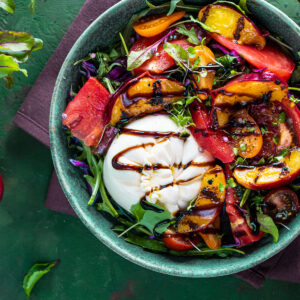 Sommersalat mit Burrata und gegrillten Nektarinen von oben in einer türkisfarbenen Keramikschale: eine ganze weiße Burrata mit dunklen Balsamico-Bändern, darum Nektarinenspalten mit Röststreifen, rote Tomatenspalten, orange und dunkle Kirschtomaten, fein gehobelter Rotkohl, Rucola und Microgreens; links daneben eine rohe halbierte Nektarine mit Stein und rot geaderte Babyblätter auf grünem Untergrund