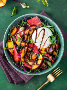 Sommersalat mit Burrata und gegrillten Nektarinen von oben in einer türkisfarbenen Keramikschale: eine ganze weiße Burrata etwas aus der Mitte nach rechts oben gesetzt, mit dunkler Balsamico-Creme beträufelt, darum Nektarinenspalten mit dunklen Röststreifen, rote Tomatenspalten, orange und dunkle Kirschtomaten, fein gehobelter Rotkohl, Rucola und Microgreens; oben links eine gegrillte Nektarinenhälfte und rot geaderte Babyblätter auf dem Tisch, daneben eine pflaumenfarbene Leinenserviette und eine goldene Gabel auf grünem Untergrund