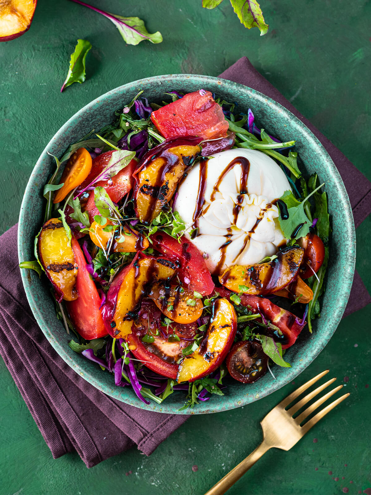 Sommersalat mit Burrata und gegrillten Nektarinen von oben in einer türkisfarbenen Keramikschale: eine ganze weiße Burrata etwas aus der Mitte nach rechts oben gesetzt, mit dunkler Balsamico-Creme beträufelt, darum Nektarinenspalten mit dunklen Röststreifen, rote Tomatenspalten, orange und dunkle Kirschtomaten, fein gehobelter Rotkohl, Rucola und Microgreens; oben links eine gegrillte Nektarinenhälfte und rot geaderte Babyblätter auf dem Tisch, daneben eine pflaumenfarbene Leinenserviette und eine goldene Gabel auf grünem Untergrund