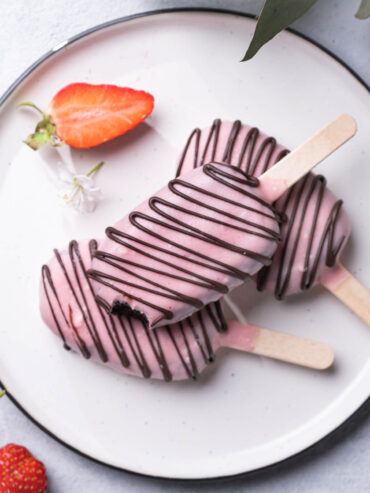 Schoko-Cakesicles am Stiel mit rosa Glasur und dunklen Schokoladenstreifen auf einem Teller mit Erdbeeren
