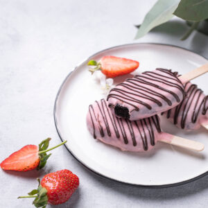 Drei Schoko-Cakesicles am Stiel mit rosa Glasur und Schokoladenstreifen auf einem weißen Teller, angerichtet mit frischen Erdbeeren