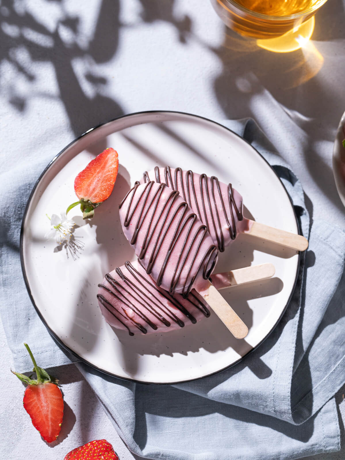 Zwei Schoko-Cakesicles am Stiel mit rosa Glasur auf einem hellen Teller, daneben ein Glas Tee