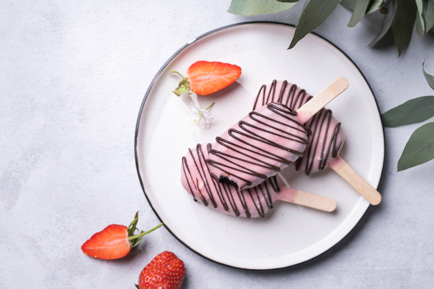 Schoko-Cakesicles am Stiel mit rosa Glasur und dunklen Schokoladenstreifen von oben auf einem Teller mit Erdbeeren
