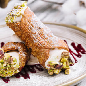 Zwei kurze, dickbauchige Cannoli auf einem hellen Steingutteller mit Goldrand: knusprig frittierte, stark blasige Teigröhren mit Puderzucker, an den offenen Enden glatte weiße Ricottacreme, dick besetzt mit grob gehackten Pistazien, daneben dunkelrote Tropfen und Bahnen Beerenspiegel auf dem Teller