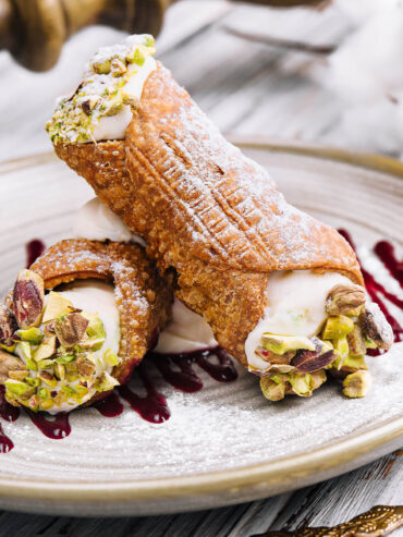 Zwei kurze, dickbauchige Cannoli übereinander auf einem hellen Steingutteller: stark blasige, goldbraun frittierte Schalen mit Puderzucker, an den weiten offenen Enden glatte weiße Ricottacreme, dick besetzt mit grob gehackten Pistazien, dazu dunkelrote Beerentropfen; im Hintergrund Baumwollzweige auf weiß gestrichenem Holz