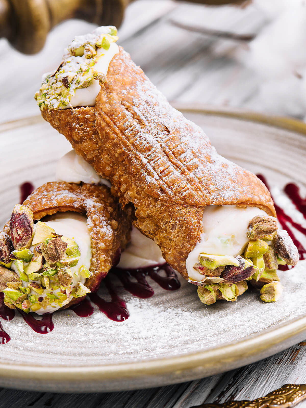 Zwei kurze, dickbauchige Cannoli übereinander auf einem hellen Steingutteller: stark blasige, goldbraun frittierte Schalen mit Puderzucker, an den weiten offenen Enden glatte weiße Ricottacreme, dick besetzt mit grob gehackten Pistazien, dazu dunkelrote Beerentropfen; im Hintergrund Baumwollzweige auf weiß gestrichenem Holz
