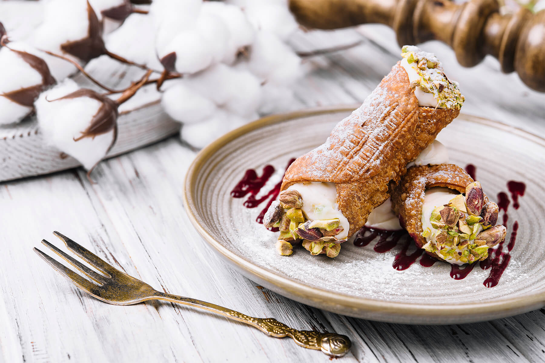 Kurze, dickbauchige Cannoli auf einem Steingutteller mit Goldrand, dahinter Baumwollzweige und eine goldene Gabel auf weiß gestrichenem Holz; die Schalen sind mit Puderzucker bestäubt, an den offenen Enden weiße Ricottacreme mit grob gehackten Pistazien, auf dem Teller dunkelrote Beerentropfen