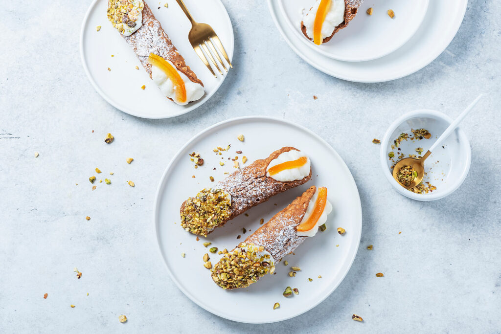 Große Cannoli Siciliani von oben auf weißen Tellern: knusprig frittierte, blasige Teigröhren dick mit Puderzucker bestäubt, ein Ende in grob gehackte Pistazien gedrückt, am anderen Ende weiße Ricottacreme mit einem breiten Stück kandierter Orangenschale; daneben eine goldene Gabel, ein Schälchen mit gehackten Pistazien und verstreute Pistazienstücke auf hellem Untergrund