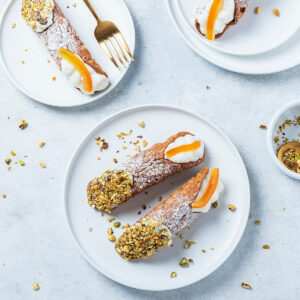 Große Cannoli Siciliani von oben auf weißen Tellern: knusprig frittierte, blasige Teigröhren dick mit Puderzucker bestäubt, ein Ende in grob gehackte Pistazien gedrückt, am anderen Ende weiße Ricottacreme mit einem breiten Stück kandierter Orangenschale; daneben eine goldene Gabel, ein Schälchen mit gehackten Pistazien und verstreute Pistazienstücke auf hellem Untergrund