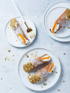 Fertige große Cannoli Siciliani von oben auf drei weißen Tellern: lange, blasig frittierte Teigröhren mit Puderzucker, ein Ende dick in grob gehackte Pistazien gedrückt, am anderen Ende weiße Ricottacreme mit einem breiten Stück kandierter Orangenschale; daneben eine goldene Gabel und verstreute Pistazienstücke auf hellgrauem Untergrund