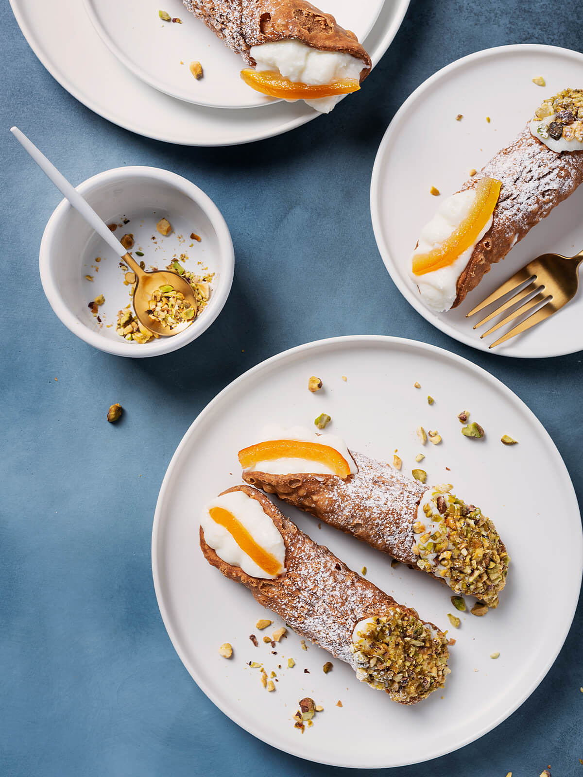 Große Cannoli Siciliani von oben auf weißen Tellern vor blauem Untergrund: knusprig frittierte, blasige Teigröhren mit Puderzucker, ein Ende in grob gehackte Pistazien gedrückt, am anderen Ende weiße Ricottacreme mit einem breiten Stück kandierter Orangenschale; daneben eine goldene Gabel und ein weißes Schälchen mit gehackten Pistazien und goldenem Löffel