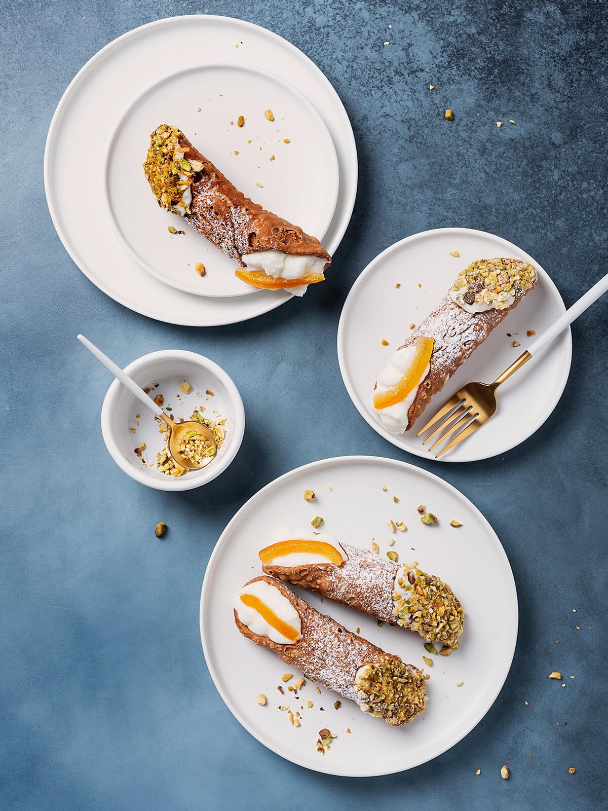 Vier große Cannoli Siciliani von oben auf weißen Tellern vor blauem Untergrund, dick mit Puderzucker bestäubt, ein Ende jeweils in grob gehackte Pistazien gedrückt, am anderen Ende weiße Ricottacreme mit einem breiten Stück kandierter Orangenschale; daneben eine goldene Gabel und ein Schälchen mit Pistazien
