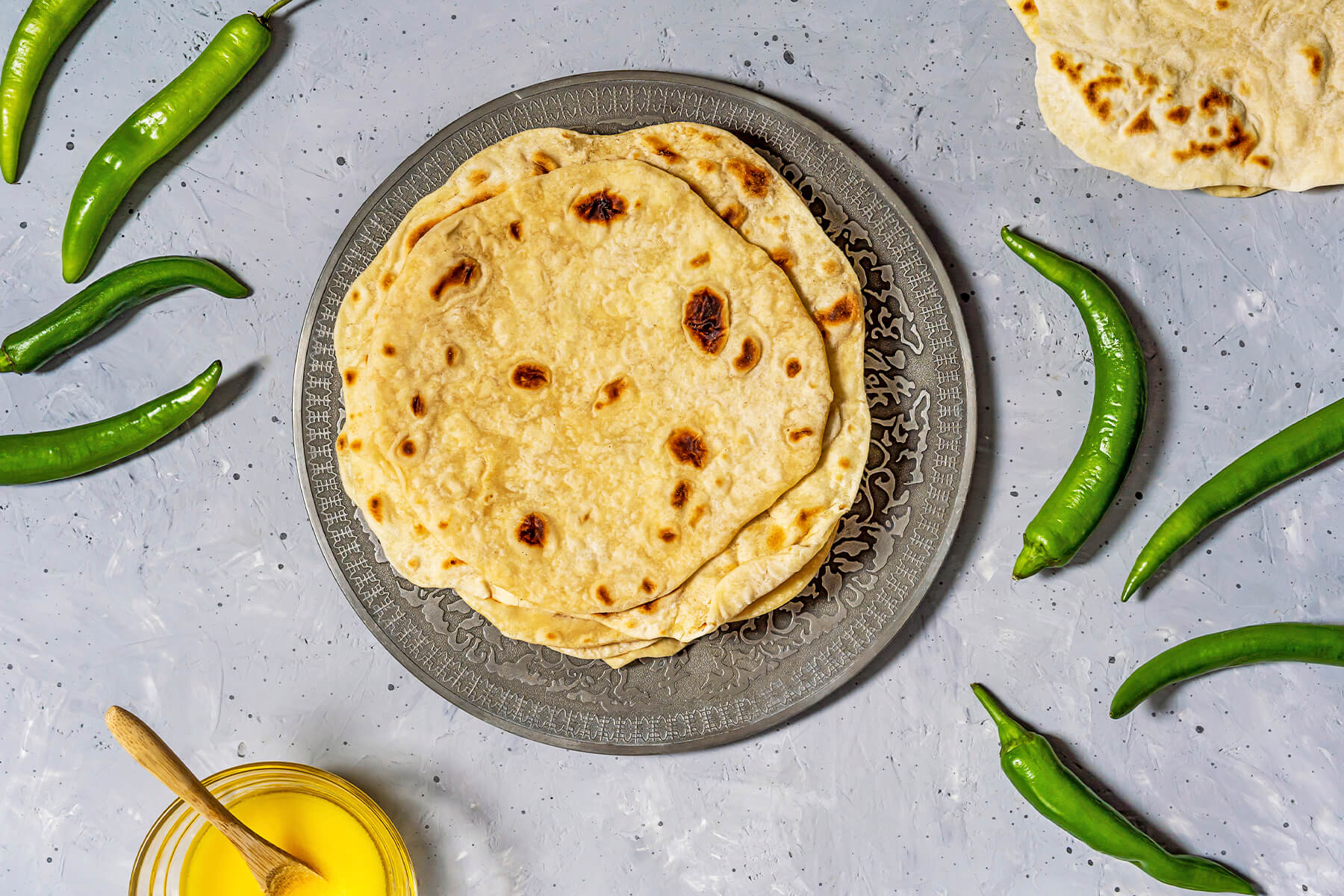 Stapel indischer Chapati-Fladenbrote von oben auf einem verzierten Teller, daneben grüne Chilis und eine Schale Ghee