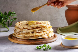 Flüssiges Ghee wird mit einem Holzlöffel über einen Stapel warmer Chapati-Fladenbrote geträufelt