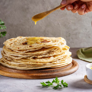 Flüssiges Ghee wird mit einem Holzlöffel über einen Stapel warmer Chapati-Fladenbrote geträufelt