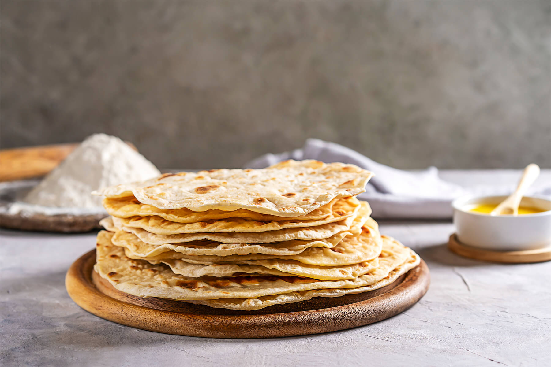 Stapel goldbrauner Chapati-Fladenbrote auf einem Holzbrett, dahinter Mehl und eine Schale Ghee
