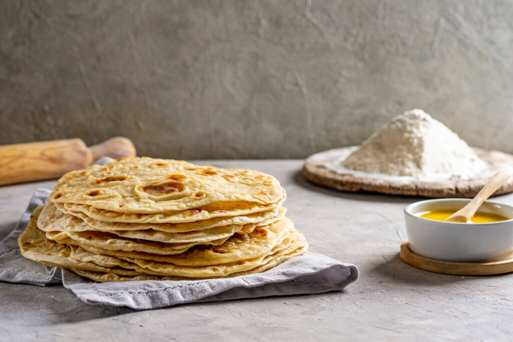 Chapati-Fladenbrote auf einem Tuch, daneben ein Nudelholz, ein Häufchen Mehl und eine Schale Ghee