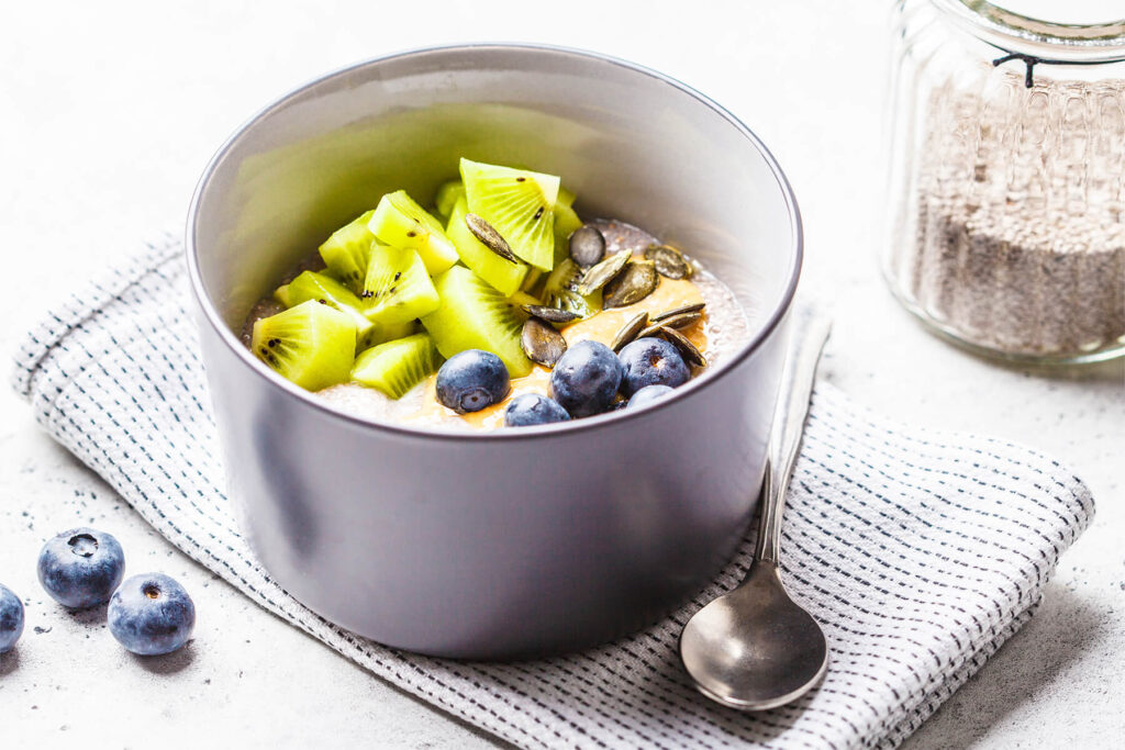 Veganer Chia-Pudding mit Kiwistücken, Blaubeeren, Erdnussmus und Kürbiskernen in einer grauen Schale, daneben ein Glas mit weißen Chiasamen