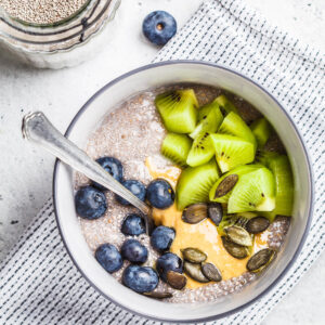 Veganer Chia-Pudding aus weißen Chiasamen mit Kiwistücken, Blaubeeren, Erdnussmus und Kürbiskernen in einer grauen Schale von oben