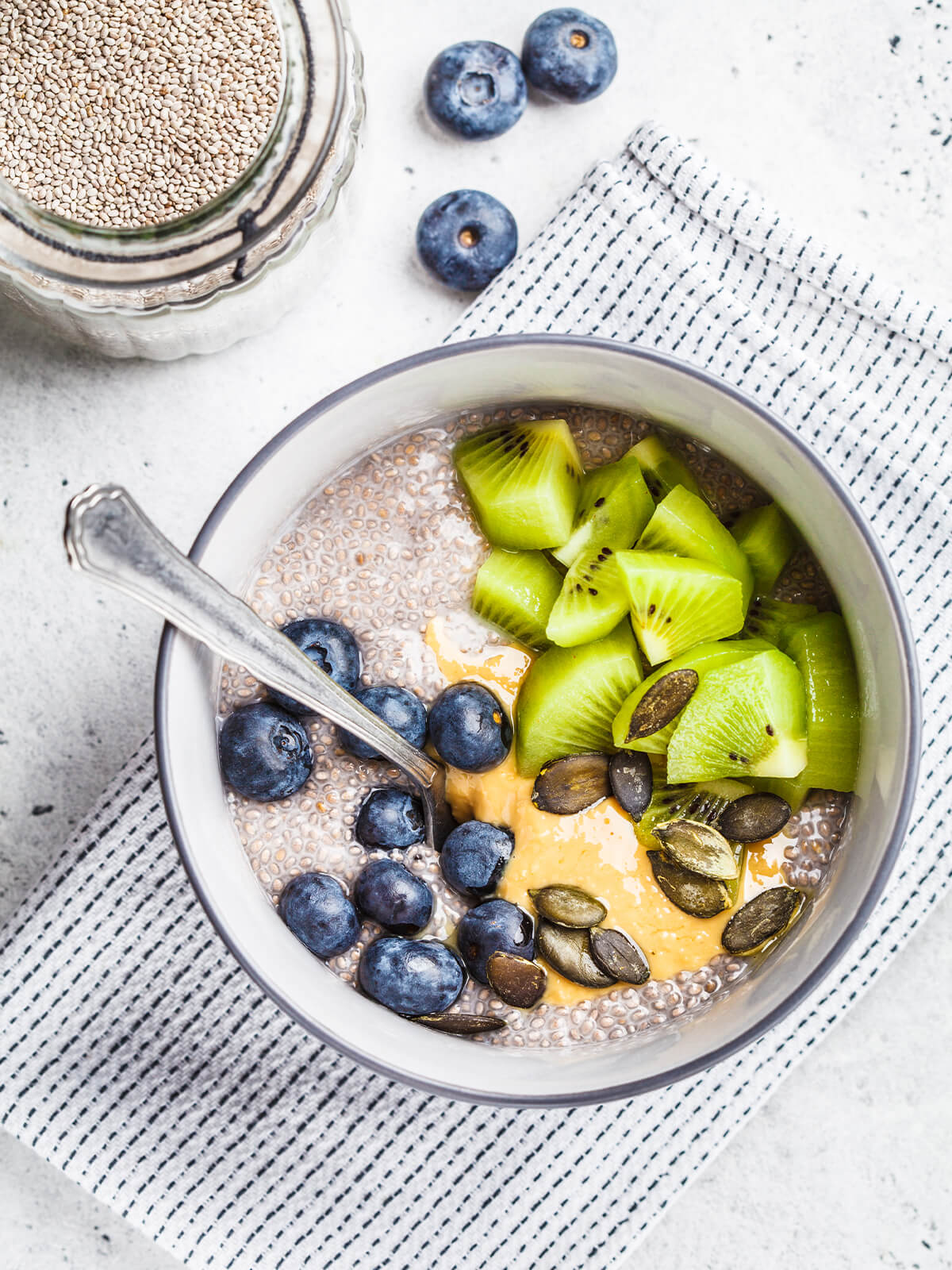 Veganer Chia-Pudding aus weißen Chiasamen mit Kiwistücken, Blaubeeren, Erdnussmus und Kürbiskernen in einer grauen Schale von oben