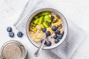 Veganer Chia-Pudding von oben mit Kiwistücken, Blaubeeren, einem Spiegel Erdnussmus, Kürbiskernen und einem Häufchen Erdnussmehl in einer grauen Schale