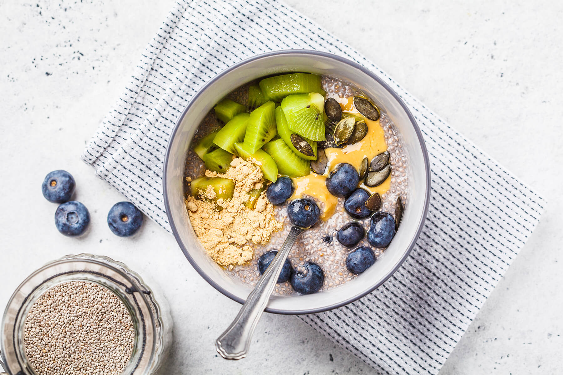Veganer Chia-Pudding von oben mit Kiwistücken, Blaubeeren, einem Spiegel Erdnussmus, Kürbiskernen und einem Häufchen Erdnussmehl in einer grauen Schale