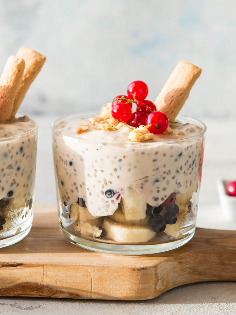 Geschichtetes Dessert in einem klaren Glas auf einem Holzbrett: unten helle Bananenscheiben mit dunklen Heidelbeeren und hellen Keksbröseln, darüber eine dicke, cremeweiße Masse voller gequollener Chiasamen, in der einzelne dunkle Heidelbeeren mitten in der Creme hängen, obenauf grobe helle Keksbrösel, eine Rispe roter Johannisbeeren und ein schräg steckendes hellbraunes Keksstäbchen; dahinter ein zweites Glas mit zwei nebeneinander schräg steckenden Keksstäbchen, im Hintergrund helles Blaugrau