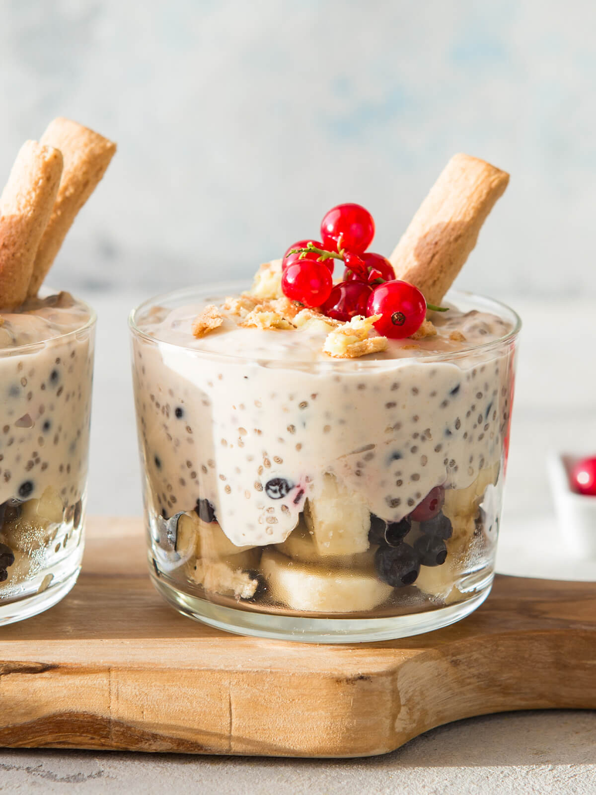 Geschichtetes Dessert in einem klaren Glas auf einem Holzbrett: unten helle Bananenscheiben mit dunklen Heidelbeeren und hellen Keksbröseln, darüber eine dicke, cremeweiße Masse voller gequollener Chiasamen, in der einzelne dunkle Heidelbeeren mitten in der Creme hängen, obenauf grobe helle Keksbrösel, eine Rispe roter Johannisbeeren und ein schräg steckendes hellbraunes Keksstäbchen; dahinter ein zweites Glas mit zwei nebeneinander schräg steckenden Keksstäbchen, im Hintergrund helles Blaugrau