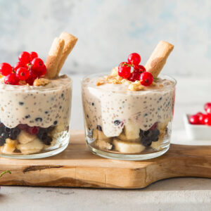 Zwei klare Gläser nebeneinander auf einem Holzbrett, beide geschichtet mit hellen Bananenscheiben und dunklen Heidelbeeren unten und dicker cremeweißer Chiacreme darüber, in der einzelne dunkle Heidelbeeren mitten in der Creme hängen; obenauf grobe helle Keksbrösel, Rispen roter Johannisbeeren und hellbraune Keksstäbchen, im linken Glas zwei nebeneinander, im rechten eines; rechts eine weiße eckige Schale mit weiteren Johannisbeeren, vorn links eine einzelne Rispe auf hellem Stein