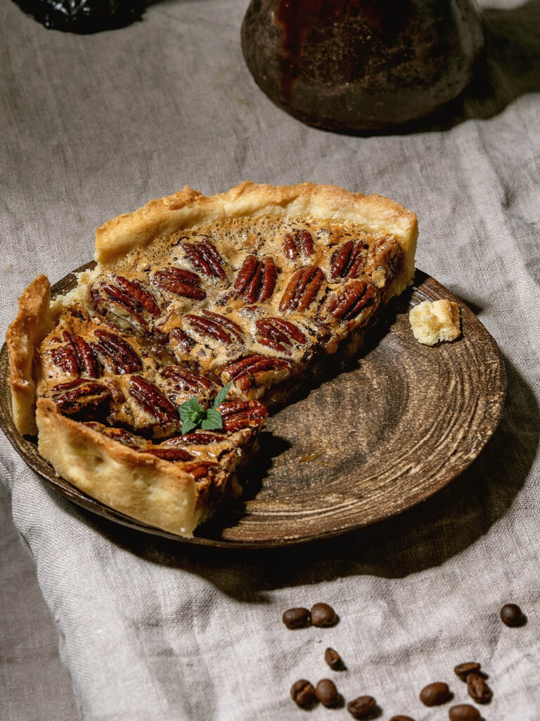 Stück klassischer Pecan Pie mit glänzender Füllung und Pekannüssen auf einem Teller, im Hintergrund Kaffee