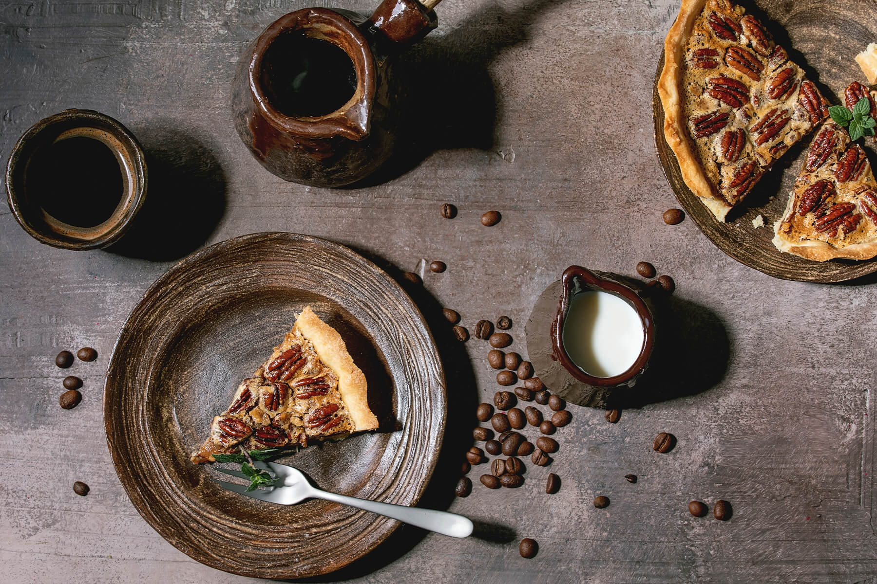 "Stück Pecan Pie auf einem Teller mit Gabel, dazu Kaffeebohnen und ein Milchkännchen auf rustikalem grau-braunem Untergrund"