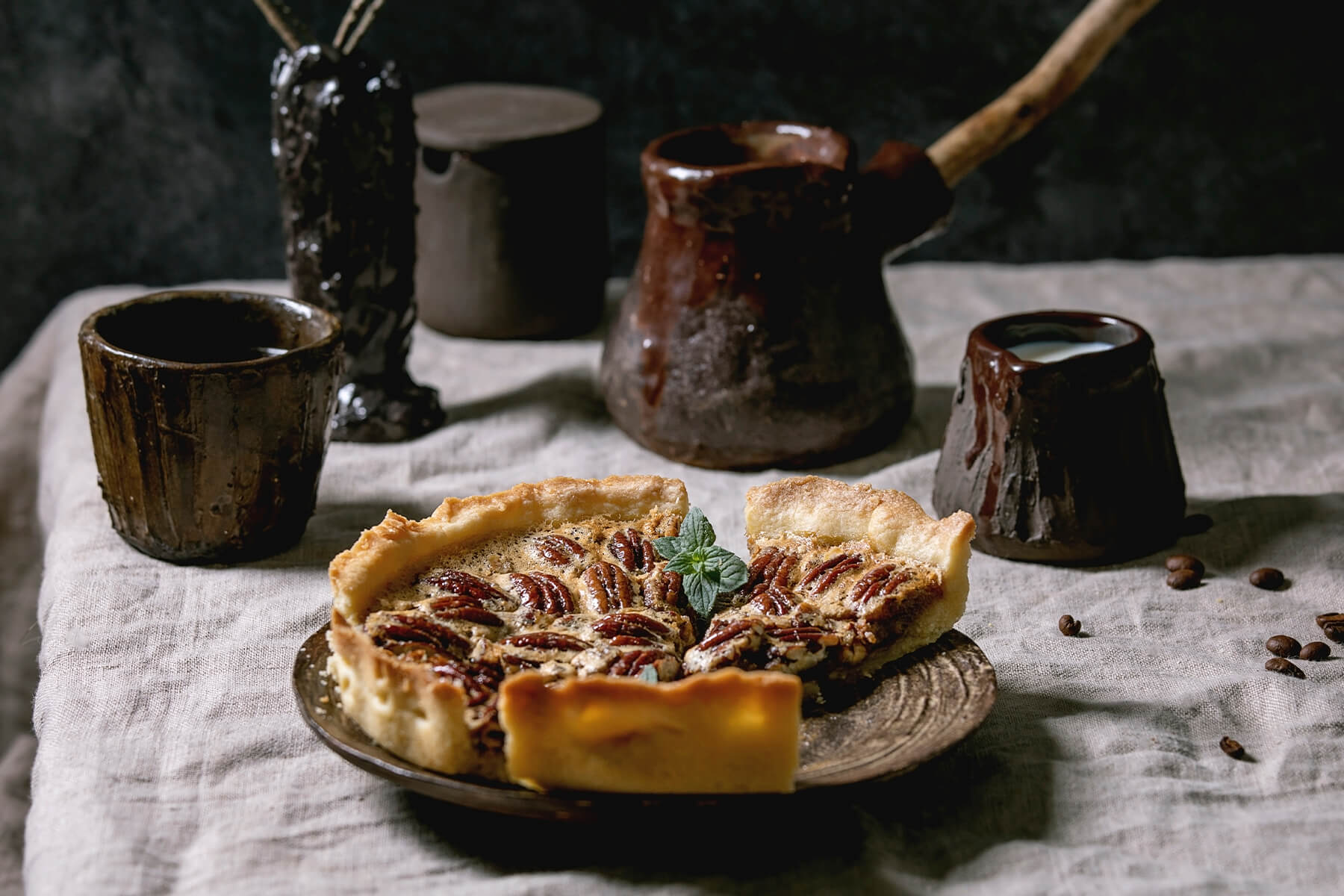 "Angeschnittener Pecan Pie mit einzelnem Stück auf rustikalem Teller, dazu Kaffeekanne und Milch in dunkler Stimmung"