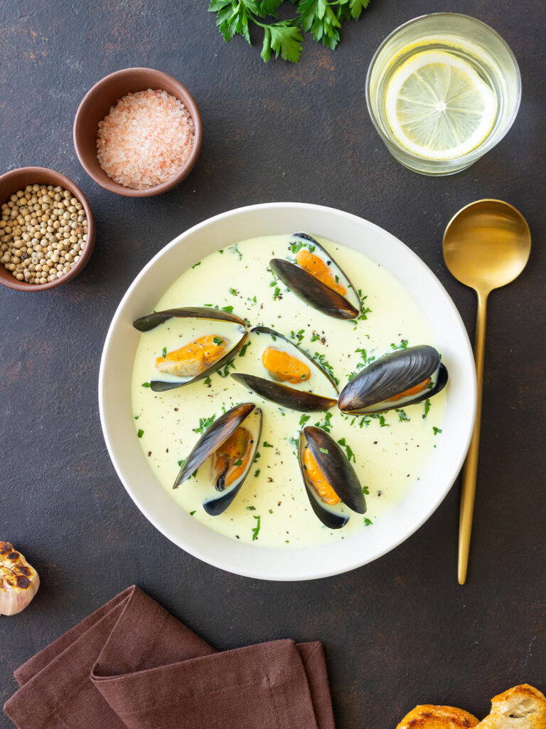 Cremige Muschelsuppe mit ganzen Miesmuscheln, Petersilie und schwarzem Pfeffer in einer weißen Schale von oben, daneben Salz, weißer Pfeffer, gerösteter Knoblauch, Zitronenwasser und geröstetes Brot