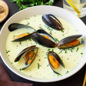 Cremige Muschelsuppe mit ganzen Miesmuscheln, Petersilie und Pfeffer in einer weißen Schale, daneben Muskatnuss, gerösteter Knoblauch und geröstetes Brot