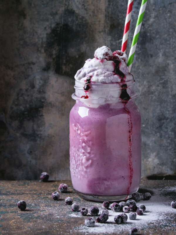 Cremiger Blaubeer-Smoothie im Glas mit Sahnehaube, Blaubeersauce und zwei Strohhalmen