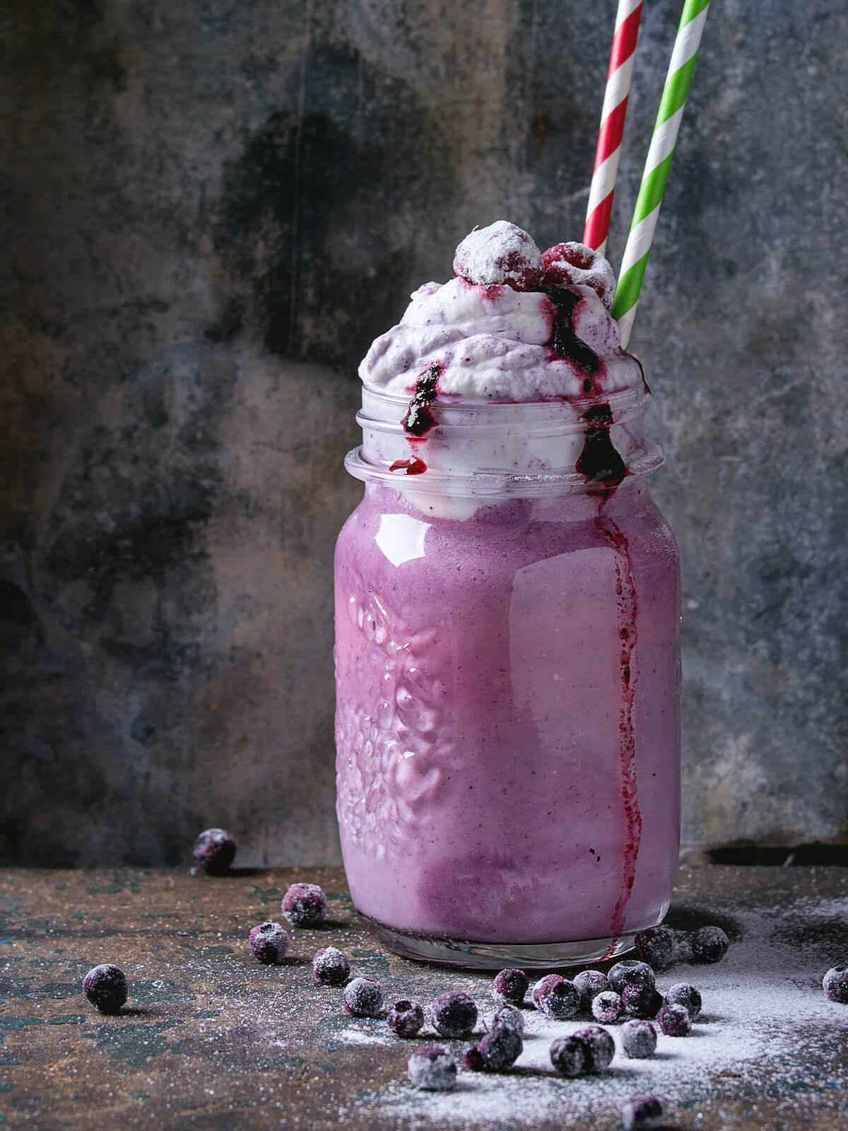 Cremiger Blaubeer-Smoothie im Glas mit Sahnehaube, Blaubeersauce und zwei Strohhalmen