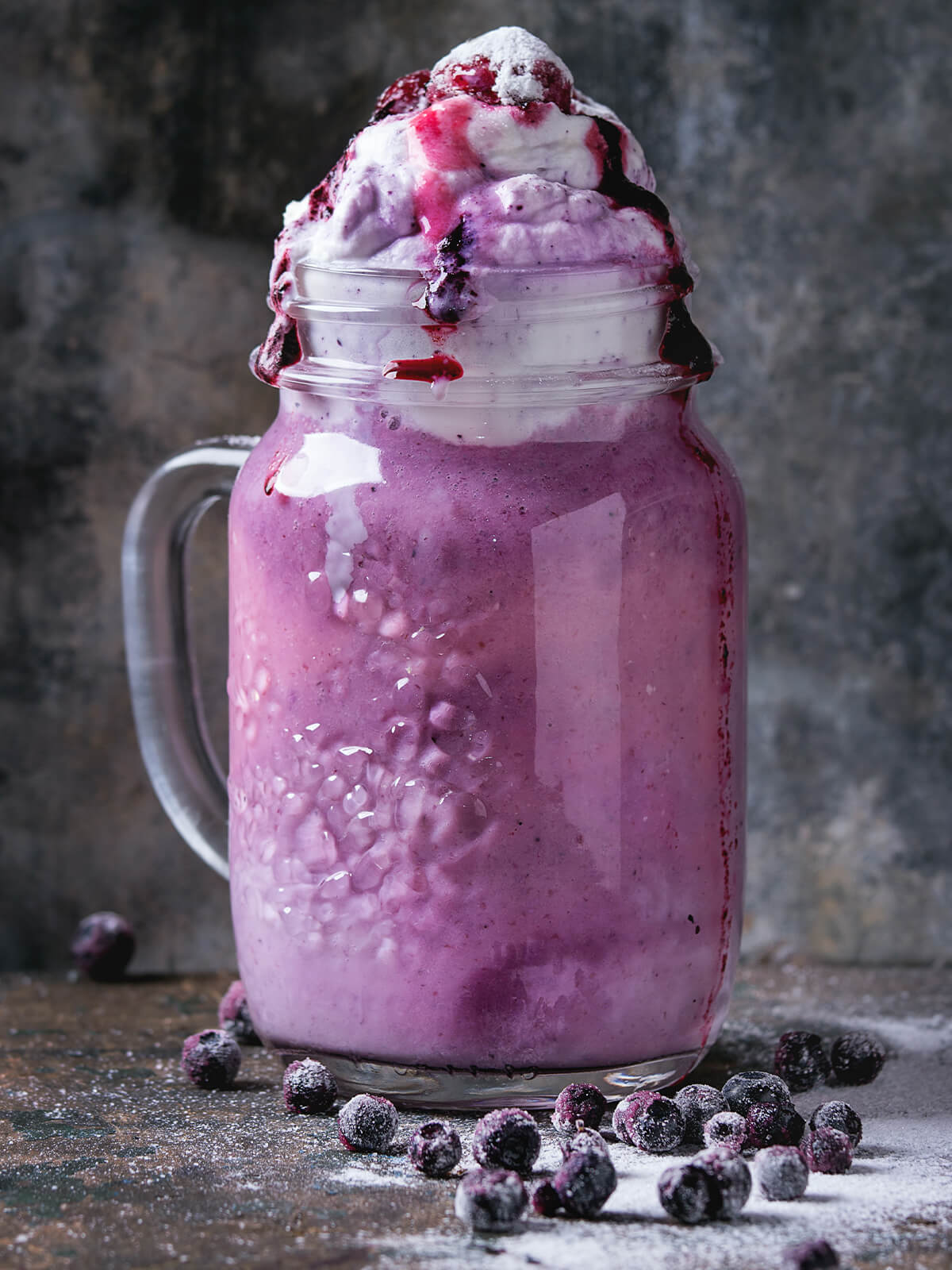 Cremiger Blaubeer-Smoothie im Henkelglas mit Sahnehaube und gefrorenen Blaubeeren, bestäubt mit Puderzucker