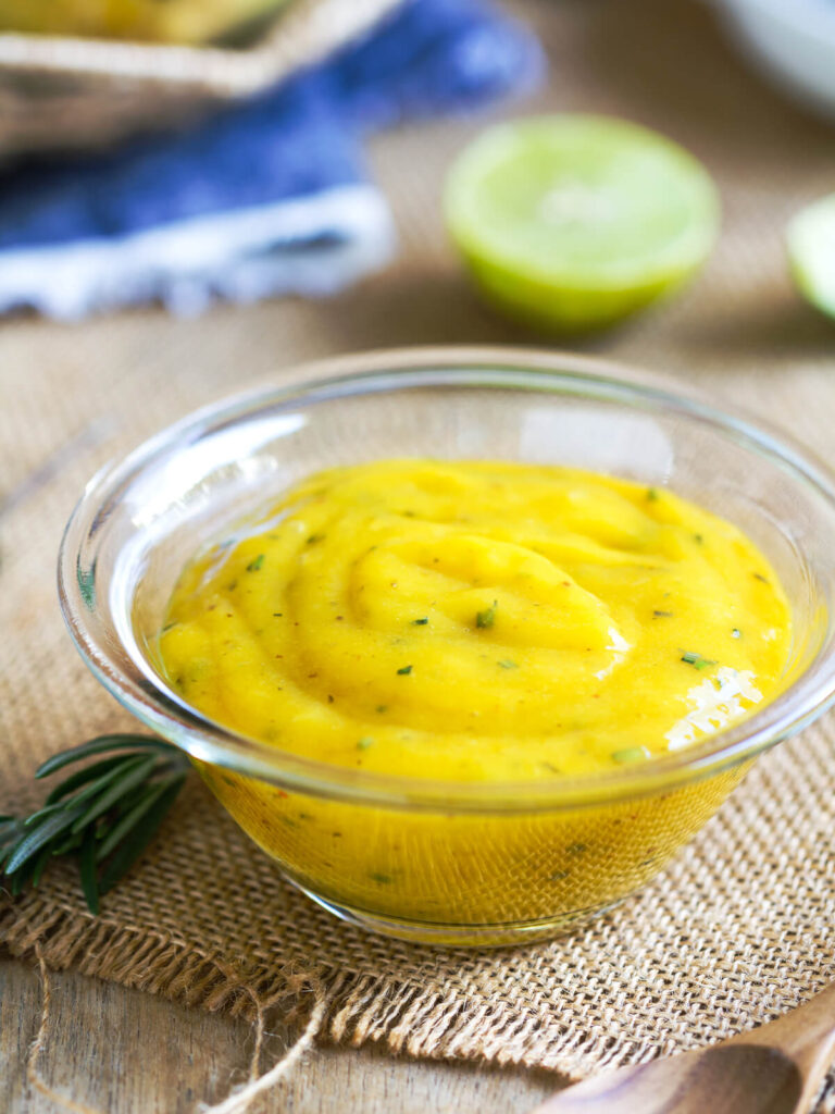 Cremiges gelbes Mango-Dressing in einer Glasschale mit Limette und einem Zweig Rosmarin auf einer Jutematte