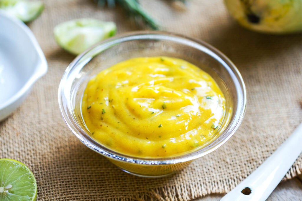 Cremiges gelbes Mango-Dressing in einer Glasschale mit Limette und Rosmarin auf einer Jutematte