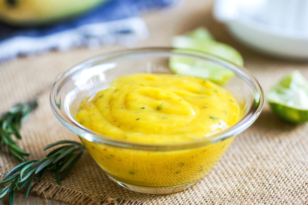 Cremiges Mango-Dressing in einer Glasschale, daneben ein Zweig frischer Rosmarin und eine halbierte Limette
