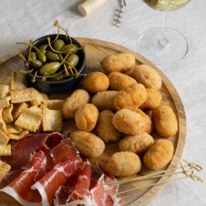 Goldbraune spanische Schinken-Kroketten auf einem Holzbrett mit Serranoschinken, Oliven, Kapern, Crackern und einem Glas Weißwein