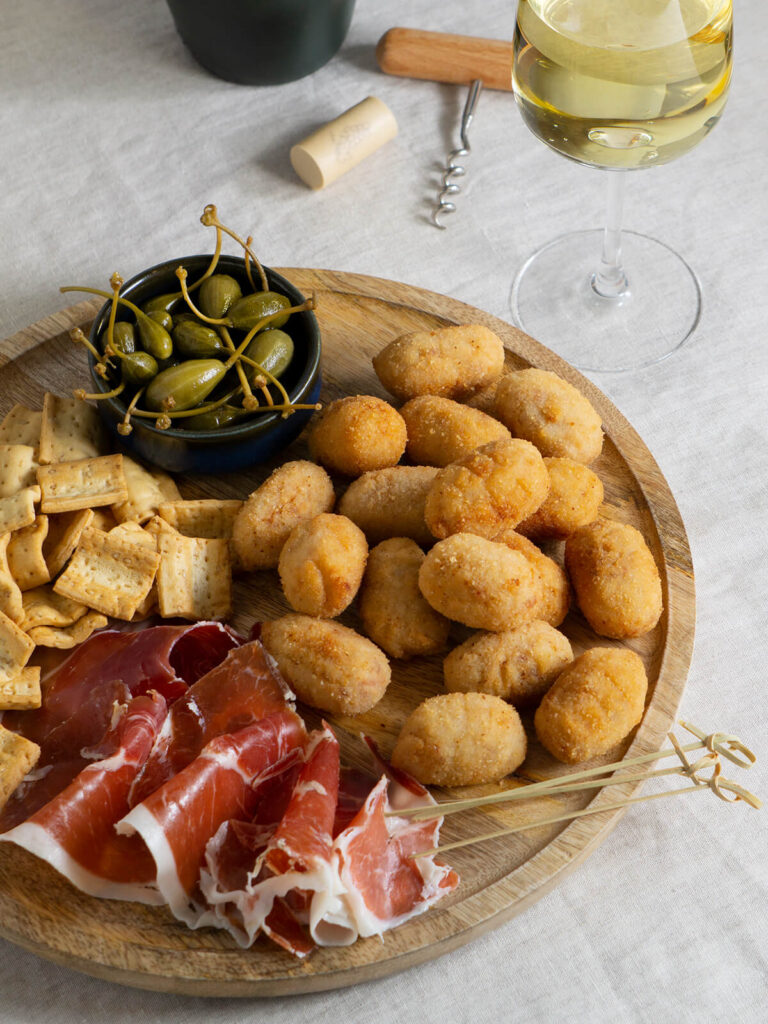 Goldbraune spanische Schinken-Kroketten auf einem Holzbrett mit Serranoschinken, Oliven, Kapern, Crackern und einem Glas Weißwein