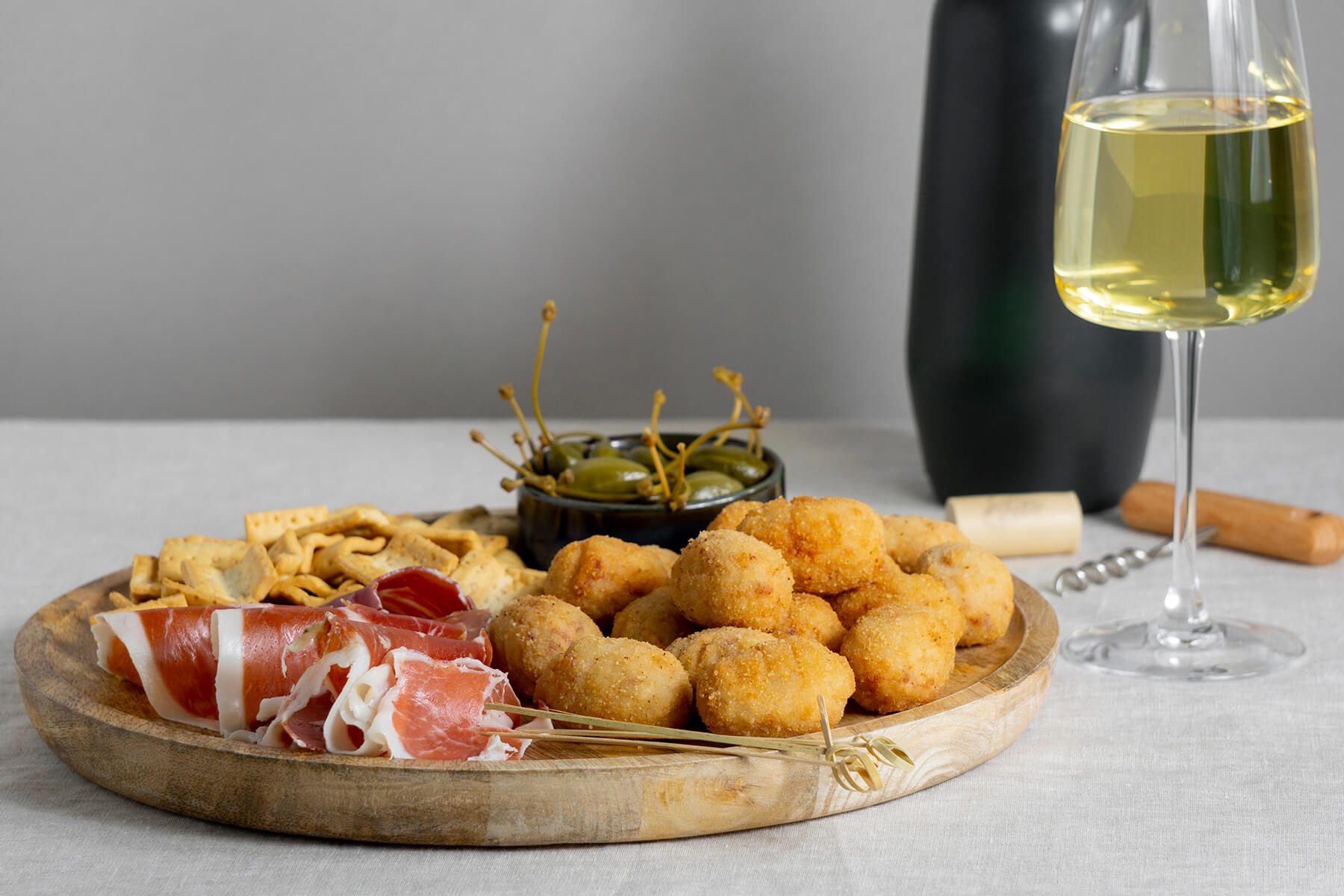 Croquetas de Jamon auf einem Holzbrett mit Serranoschinken, Oliven und Crackern, daneben ein Glas Weißwein