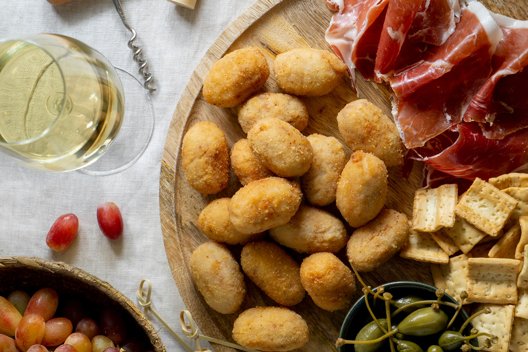 Goldbraune Croquetas de Jamon von oben auf einem Holzbrett, daneben Serranoschinken, Cracker, Kapern, Weintrauben und ein Glas Weißwein