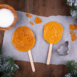 Zwei goldbraune Dalgona-Zuckerplätzchen am Stiel mit eingedrücktem Lebkuchenmann- und Tannenbaum-Muster, dazu eine Schale Zucker