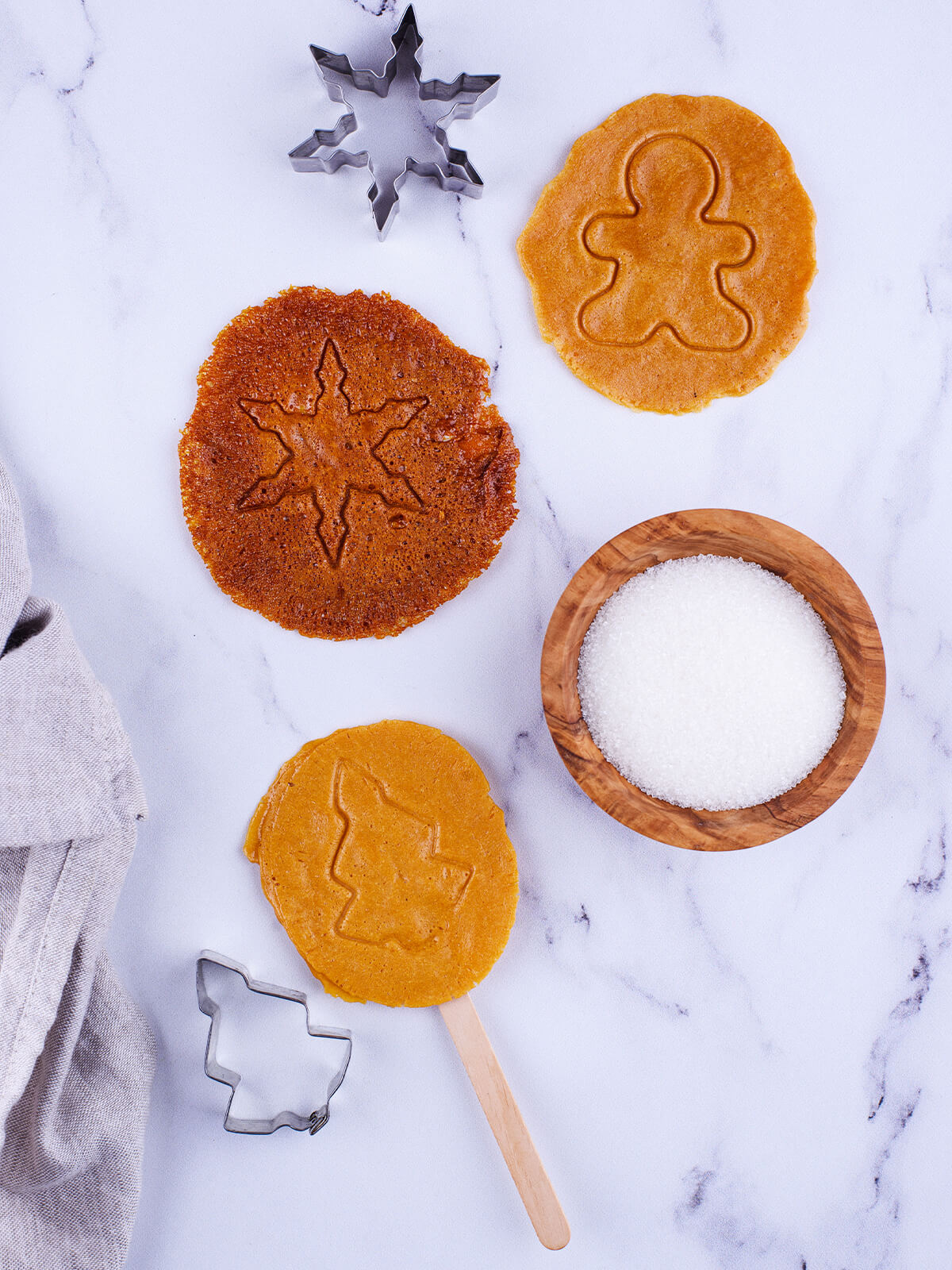 Drei Dalgona-Zuckerscheiben in verschiedenen Bräunungsgraden mit Schneeflocken-, Lebkuchen- und Tannenbaum-Muster neben Ausstechformen und einer Schale Zucker