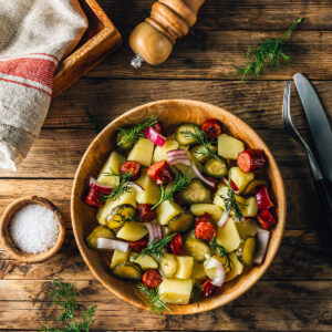 Deutscher Kartoffelsalat ohne Mayonnaise von oben in einer Holzschüssel mit Kartoffelstücken, angebratenen Scheiben geräucherter Wurst, Gewürzgurken, roten Zwiebeln und frischem Dill