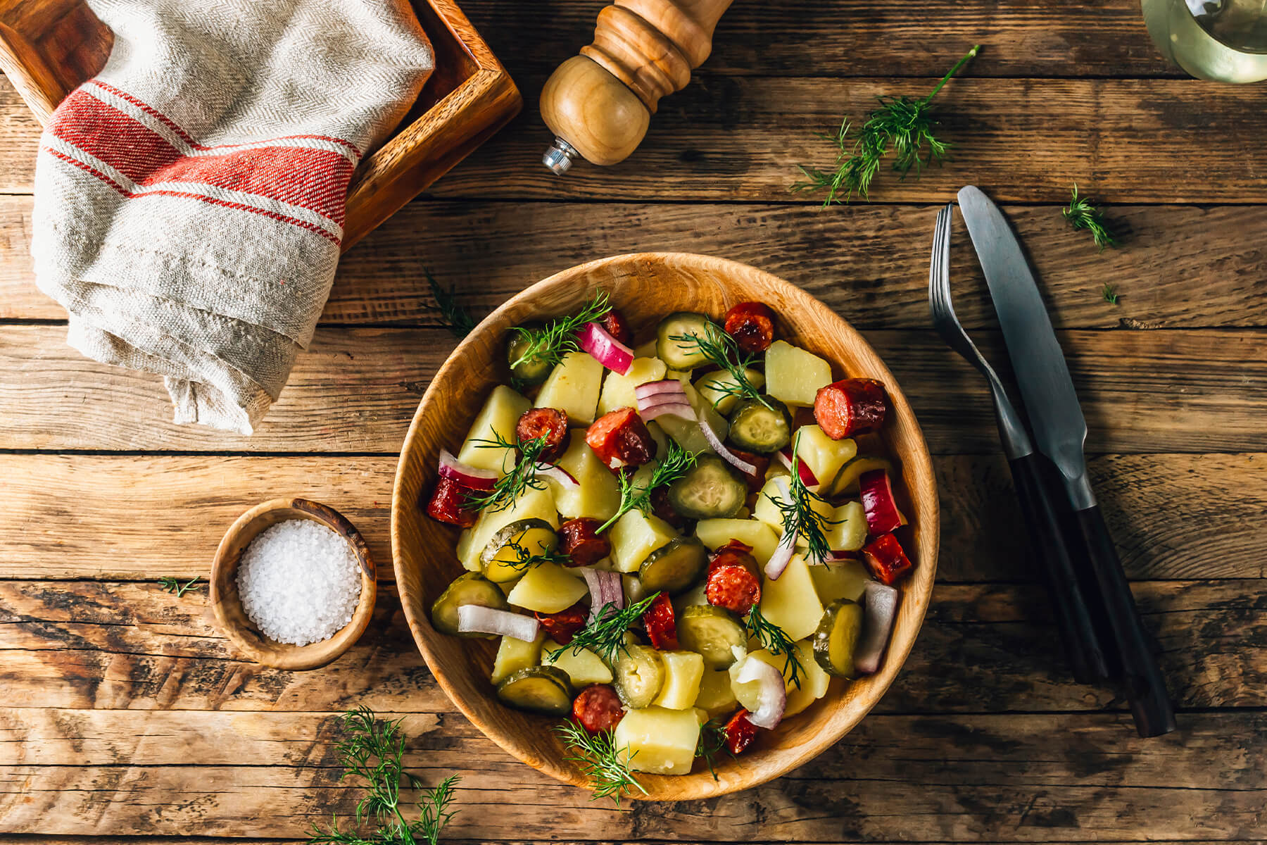 Deutscher Kartoffelsalat ohne Mayonnaise in einer Holzschüssel von oben, daneben Leinentuch, Pfeffermühle und Salz, gefüllt mit Kartoffelstücken, angebratener geräucherter Wurst, Gewürzgurken, roten Zwiebeln und Dill